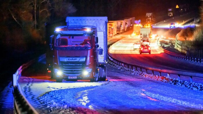 Lastbil på snöig väg nattetid med blå och röda ljusreflektioner, andra fordon syns längre fram.