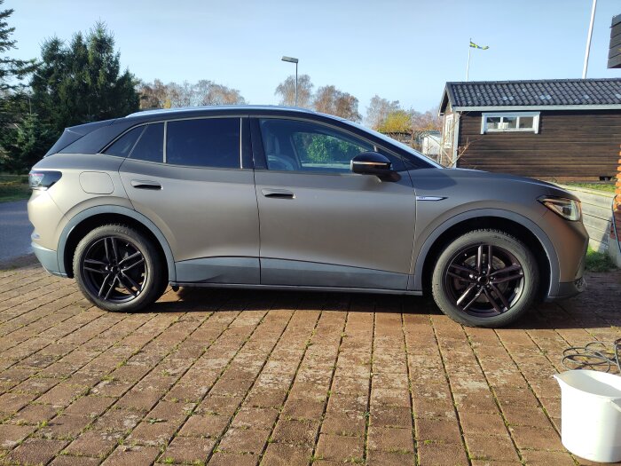 Silvergrå SUV parkerad på stenlagd uppfart, omgivning med träbyggnad och svensk flagga i bakgrunden.