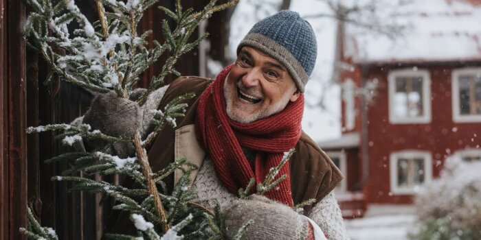 En leende person i vinterkläder håller en snöig gran framför ett rött hus, redo för julförberedelser.