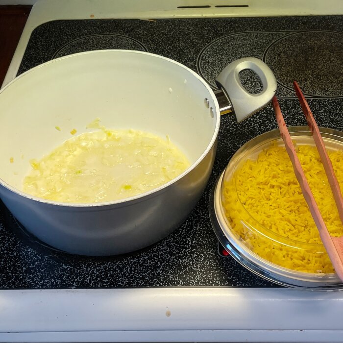 Vit gryta med lökfräs på svart spis, bredvid gul ris i plastbehållare med chopsticks ovanpå.