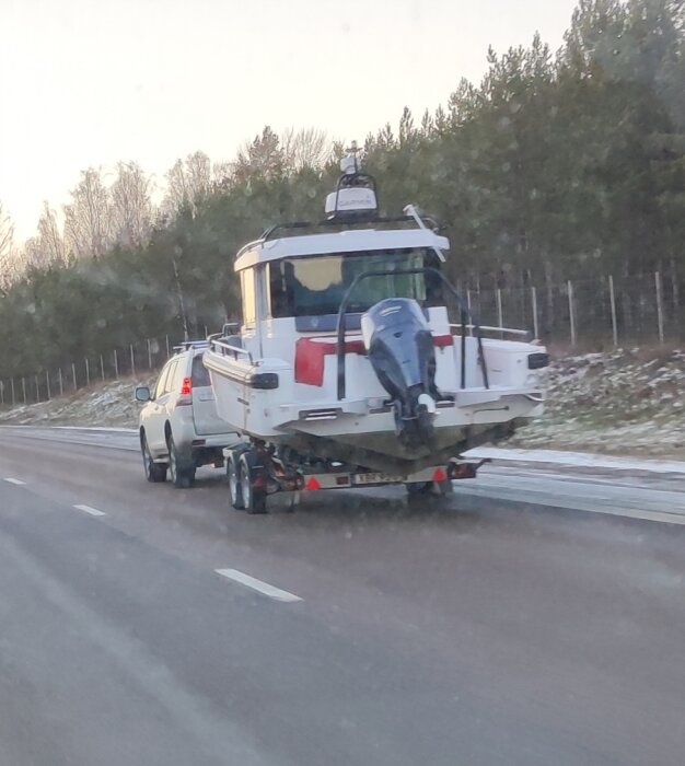 Bil drar båt på motorväg, bakhjulen nära att lyfta från marken, båtvagn saknar belysning. Skog och snöiga vägkanter syns i bakgrunden.