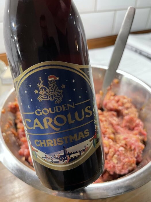 Flaska Gouden Carolus Christmas öl framför en skål med blandning för köttbullar på ett köksbord.