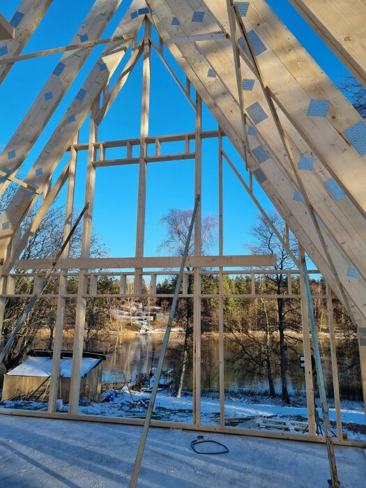 Träram av husbygget med A-formad takstomme i solig miljö. Byggplats med sjö och skog i bakgrunden.