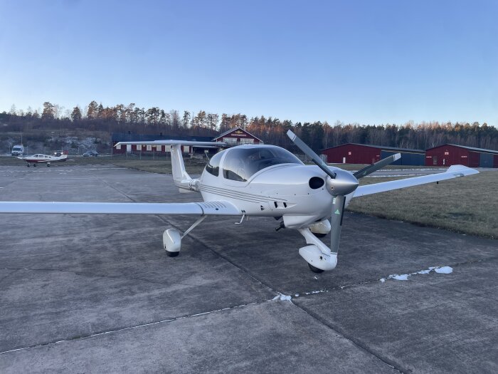 Litet flygplan står parkerat på en flygplats, med röda byggnader och skog i bakgrunden under en klarblå himmel.