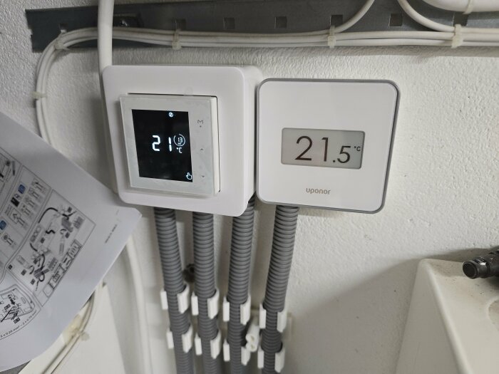 Zigbee-styrd el-termostat och trådbunden Uponor-termostat visar temperaturen 21°C respektive 21.5°C mot en vit vägg med kablar.