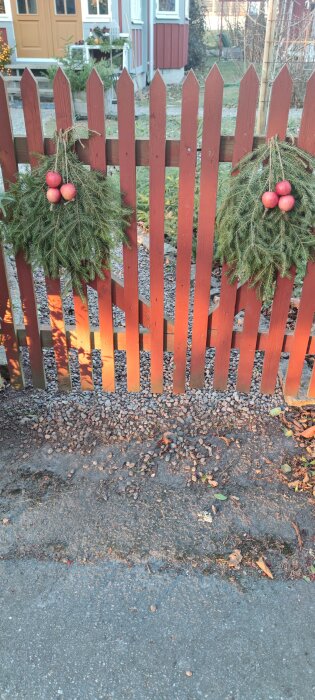 Juldekorerade granruskor med röda äpplen hänger på en brun grind framför ett hus.