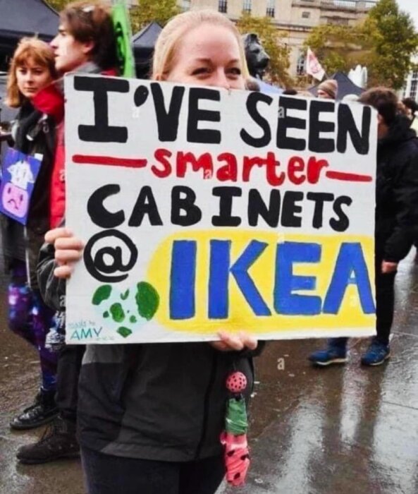 En kvinna håller en skylt under en demonstration där det står "I've seen smarter cabinets @ Ikea" i en folkmassa.