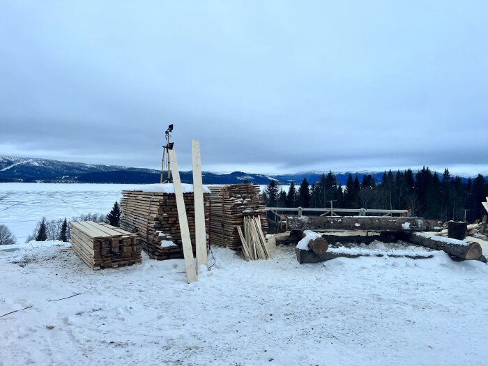 Staplar av trävirke på ett snötäckt fält med utsikt över en sjö och skog i bakgrunden.