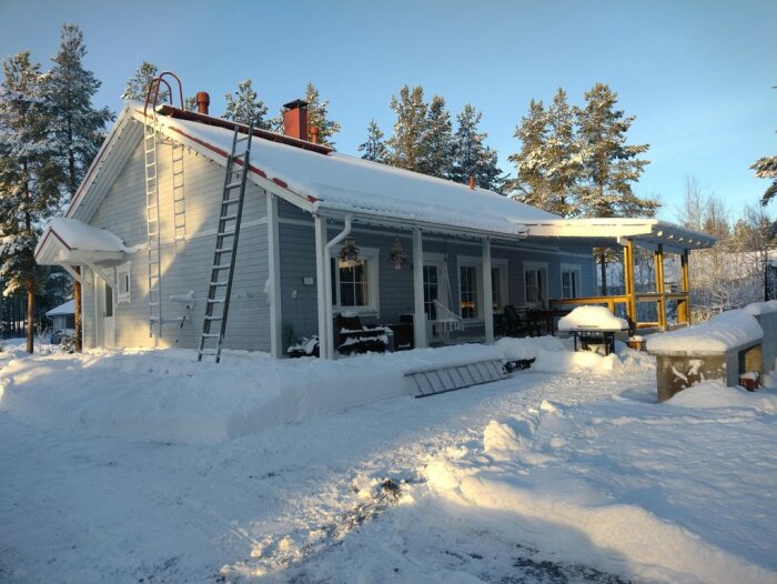 Hus med snötäckt tak och två stegar lutade mot väggen; julbelysning synlig längs taket.