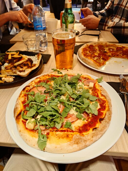 Pizza med skinka, ananas, blåmögelost och ruccola på tallrik, med ölglas och andra rätter på bordet.