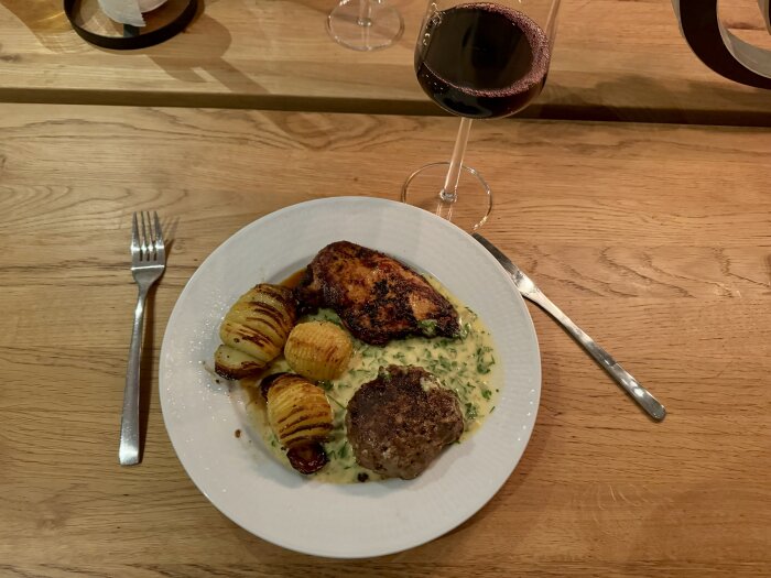 Tallrik med kycklingfilé, hasselbackspotatis och wallenbergare i brunsås på ett träbord, bredvid ett glas med röd dryck.