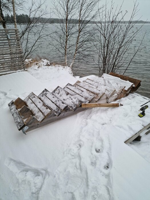 Snötäckt badtrappa vid en sjö, omgiven av vinterlandskap med träd och vatten i bakgrunden.