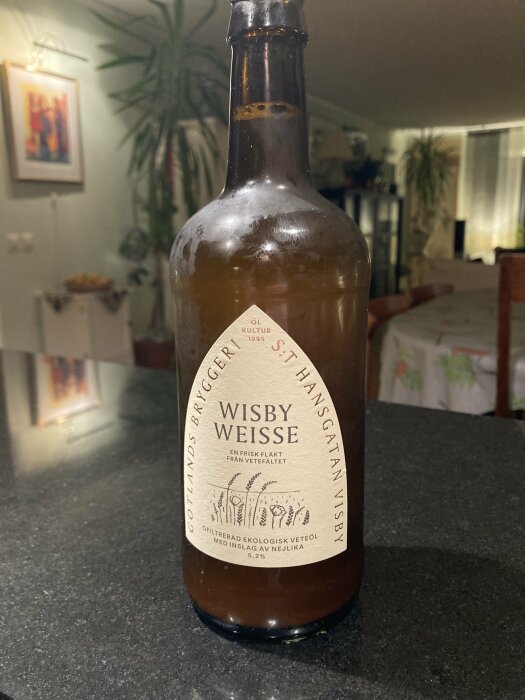 Flaska med Wisby Weisse öl från Gotlands Bryggeri, står på ett mörkt bord i ett vardagsrum med växter och konst på väggarna.