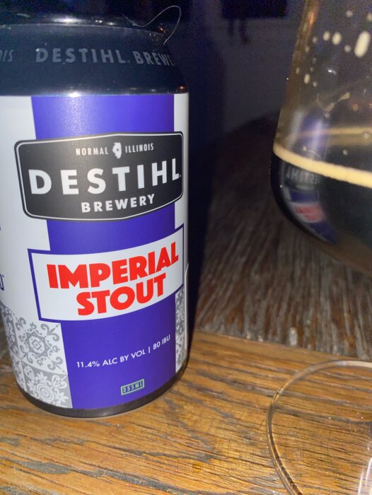 En burk Destihl Brewery Imperial Stout på ett träbord bredvid ett halvt fyllt glas med öl.