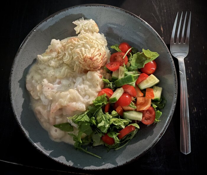 Tallrik med fiskgratäng, mos och sallad, dekorerad med räkor och dillsås, serverad med citron och en gaffel vid sidan.