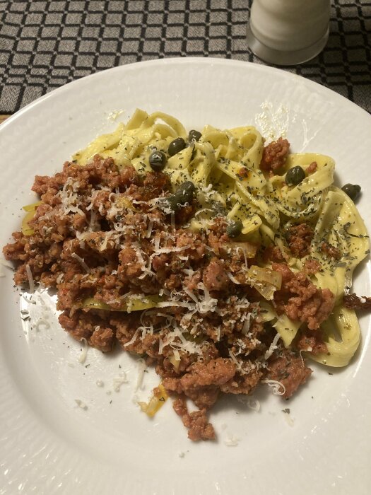 Gräddstuvad Tagliatelle med salsicciafärs, toppad med kapris, basilika, parmesan och citronzest på en vit tallrik.