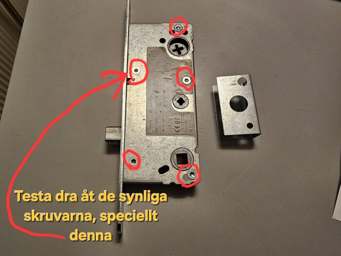 ASSA 2002 låshus med röda markeringar för justering av skruvar, text: "Testa dra åt de synliga skruvarna, speciellt denna".