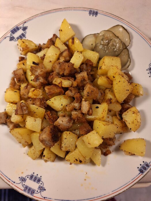 Hemlagad pytt i panna med potatis, lök och schnitzelbitar på en tallrik, serverad med inlagd gurka.