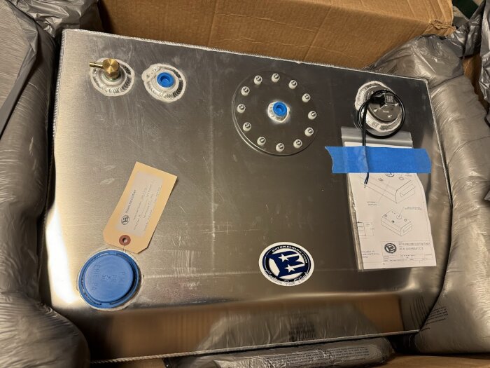 Bränsletank i metall med anslutningar, förberedd för EFI, placerad i en kartong. Innehåller etiketter och dokumentation, delvis täckt av blå tejp.