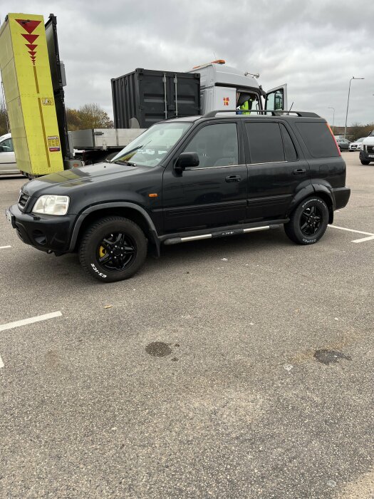 Svart SUV parkerad på asfalterad yta, omgiven av andra fordon, med en lastbil i bakgrunden under en mulen dag.