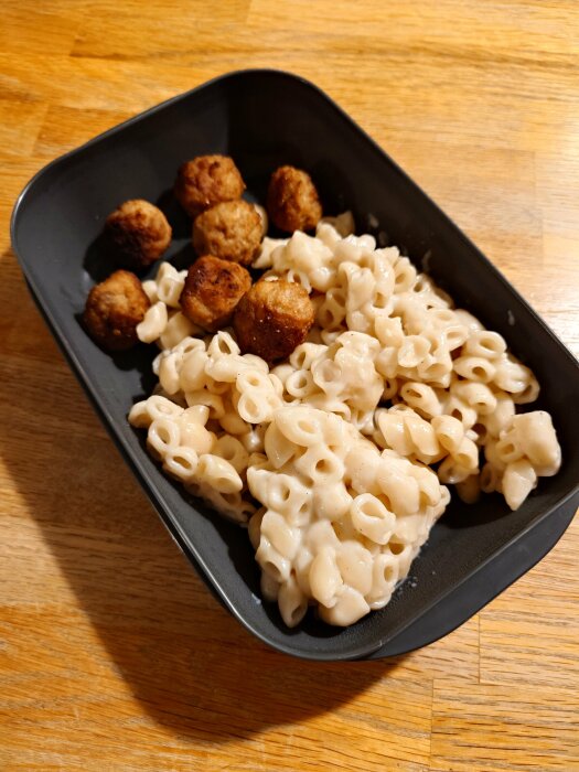 Svart matlåda med stuvade makaroner och köttbullar på ett träbord.