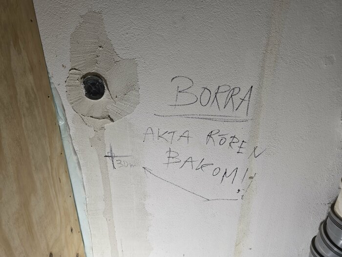 Vägg med borrhål och texten "BORRA" samt "AKTA RÖREN BAKOM!" för håltagning till vattenrör. Plywood och ledningar syns bredvid.