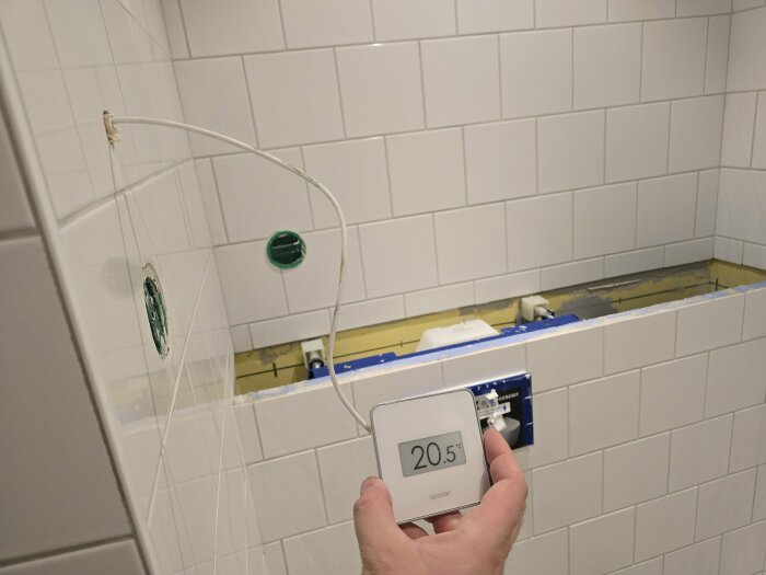 Hand holding a thermostat set to 20.5°C against tiled bathroom wall with visible plumbing installation.