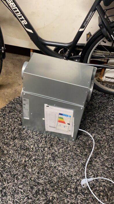 Ventilationsaggregat på en matta bredvid en cykel, med en energiklassetikett synlig framtill.