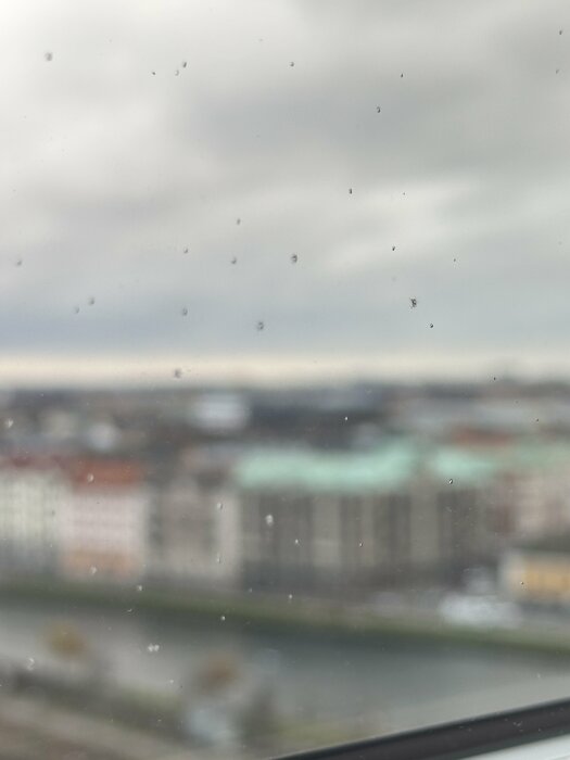 Närbild av fönsterglas med små prickar liknande stenskott, mot en suddig bakgrund av byggnader och himmel.