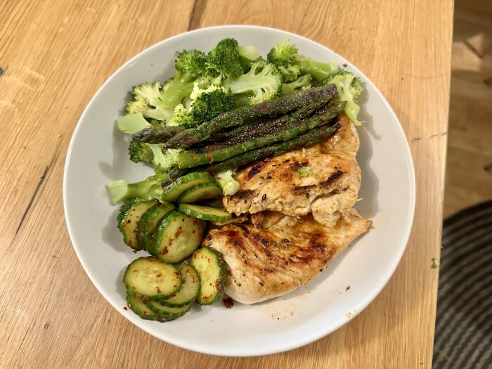 Tallrik med grillad kyckling, sparris, broccoli och gurkskivor på träbord.