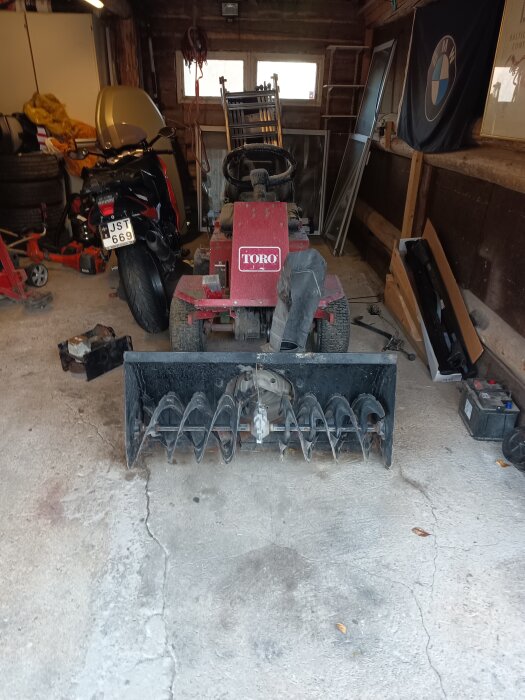 Toro åkgräsklippare i ett garage, med en snöslunga framtill. Verktyg och motorcykel syns i bakgrunden.