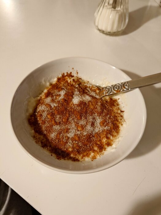 En tallrik risgrynsgröt med kanel och socker samt en sked på ett bord, bredvid en sockerströare.
