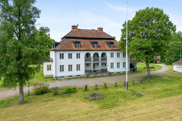 Stor vit villa med rött tegeltak, omgiven av grönska och träd, sedd från en gräsbevuxen uppfart på Tidstrandsvägen i Falu kommun.