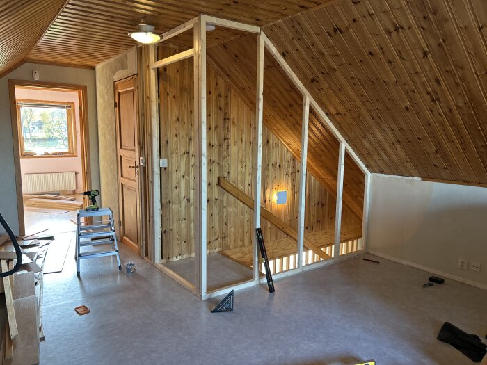Rum under renovering med träpaneltak, öppning mot trappa. En stege med verktyg syns, processen med att bygga väggar pågår.