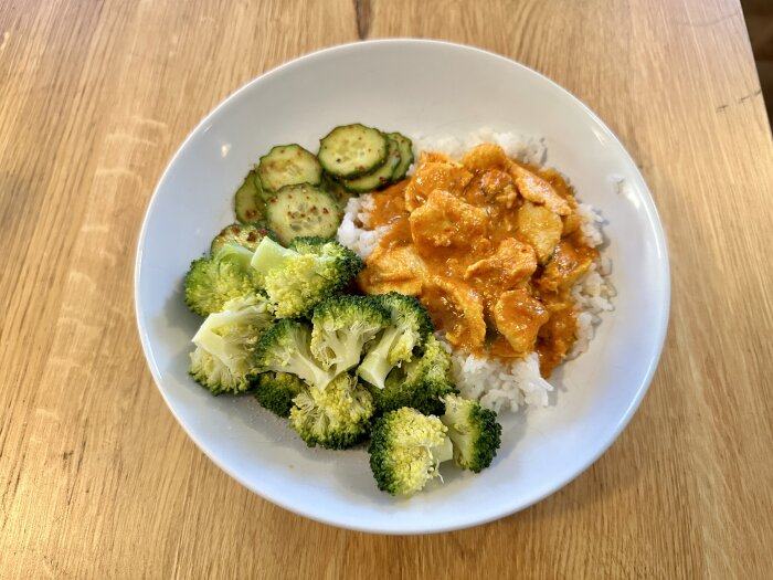 Färdigrätt med panang curry kyckling, ris, skivad gurka och ångkokt broccoli i en skål på ett träbord.