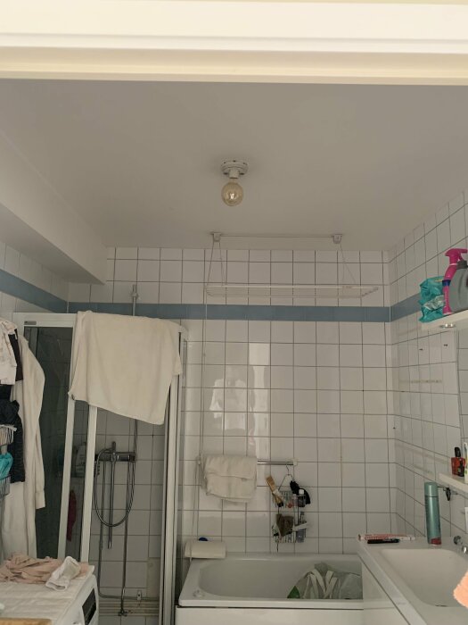 Före renovering av badrum med vitt kakel, duschhörna och badkar. Lösa kläder och artiklar syns på hyllor och tvättmaskin.