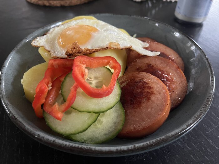 Tallrik med stekt ägg, korvskivor, potatis, gurka och paprikaringar.