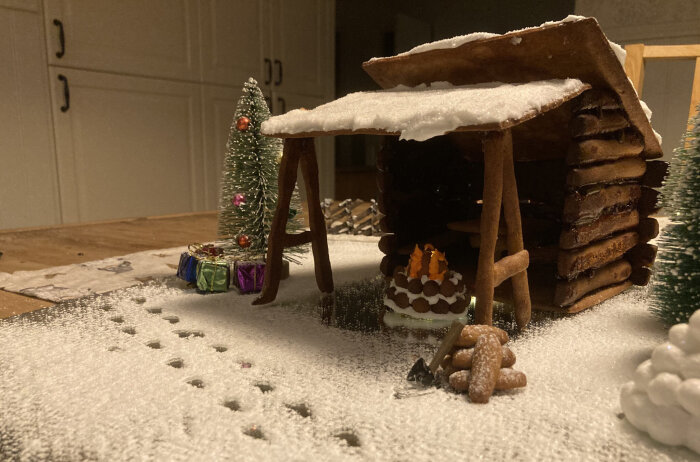 Gingerbread house with snowy roof, surrounded by a small Christmas tree, colorful gifts, and a mini campfire.