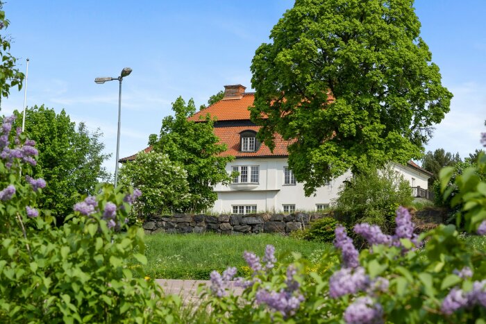 Lyktstolpe mitt i en trädgård med ett hus i bakgrunden och lila blommor i förgrunden.