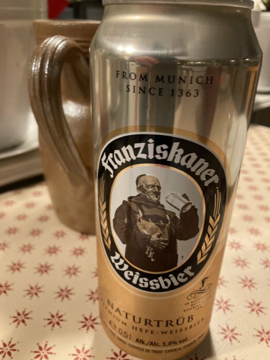 En ölburk med Franziskaner Weissbier står på ett bord med mönstrad duk, i bakgrunden syns ett keramiskt ölkrus.