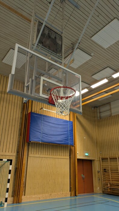 Basketkorg i idrottshall med träpanel och blå madrass på väggen.