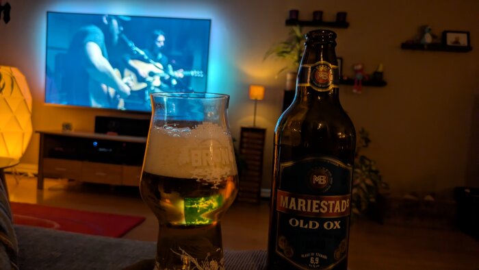 En Mariestads Old Ox ölflaska och ett glas öl framför en TV med livemusik.