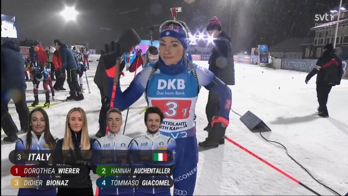 Skidåkare i tävlingsdräkt står på snötäckt arena efter en tävling, omgiven av andra deltagare och tränare. Texten "DKB Das kann Bank" synlig på dräkten.