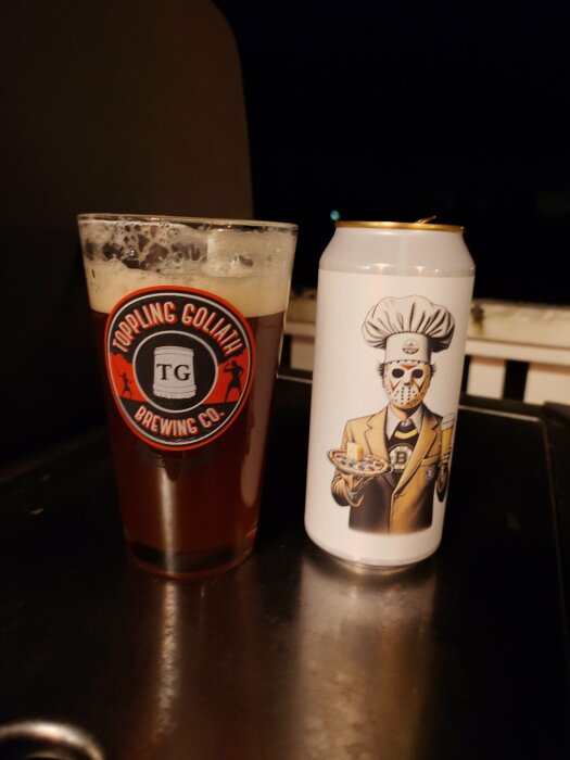 Ett glas med mörk IPA från Toppling Goliath och en ölburk med en maskerad kock på etiketten, stående på ett bord.