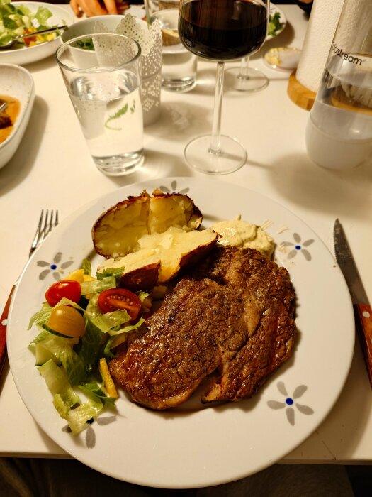 Tallrik med grillad biff, entrecote, bakad potatis, sallad och ett glas rött vin på ett dukat bord.