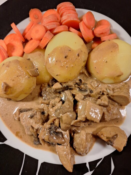 Tallrik med kycklinggryta, potatis och morotsbitar. Grytan innehåller svamp och serveras med krämig sås.