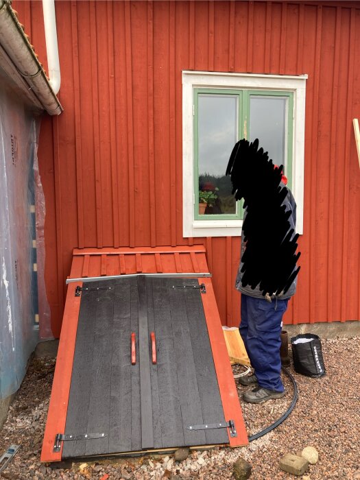 Rött träpanelhus med nyrenoverad källardörr i svart och rött. Person i arbetskläder står bredvid. Grusmark med verktyg syns.