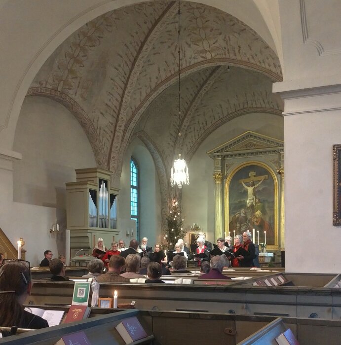 Kör sjunger i en kyrka med vackert dekorerat tak och altartavla, under en Gudstjänst inför församlingen som sitter i bänkarna.