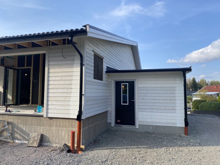 Baksidan av ett garage med en Prido-dörr, träpanel på väggen och tak med hängrännor, under en klarblå himmel.