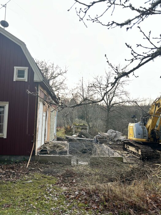 Röd träfasad med rivningsarbete, synlig grund och grävmaskin på en tomt med träd och gräsmatta i bakgrunden.